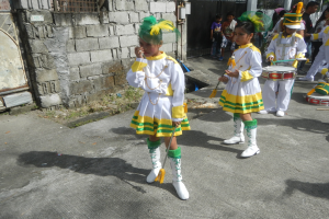 Gruppe junger Mädchen in weißen und gelben Outfits, die marschieren und Musikinstrumente spielen, vorbei an einer Wand, einem Tor und Bäumen.