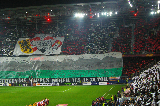 Ein Fußballstadion voller Zuschauer, einige stehen, andere sitzen, mit einer großen zentralen Fahne, perimeter-Boards und Überkopfbeleuchtung, die eine lebendige Atmosphäre schaffen.