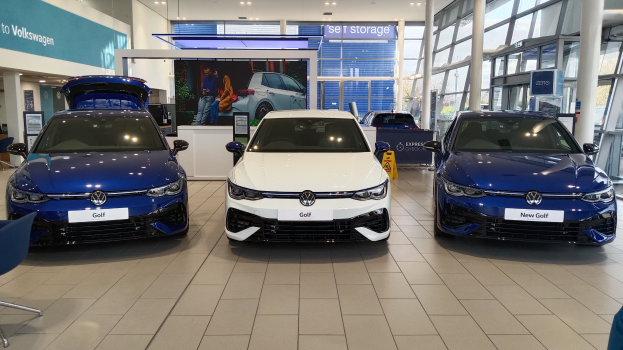 Volkswagen-Showroom mit drei blauen und weißen Autos, einem Tisch mit Gegenständen, Informationsplakaten, Glaswänden und Deckenleuchten.