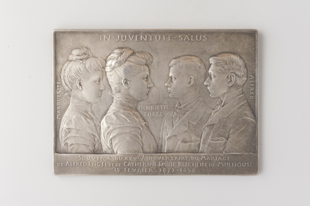 Silberne Plakette mit einem Foto von drei Personen und eingraviertem Text, vor einem weißen Hintergrund, wahrscheinlich zur Erinnerung an eine Hochzeit.