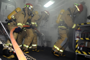 Feuerwehrleute in Schutzausrüstung arbeiten in der Nähe eines Löschfahrzeugs mit Ausrüstungsgestellen und sichtbarem Hintergrund.