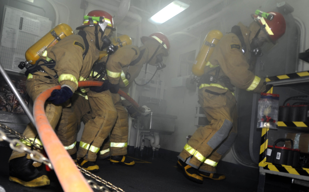 Feuerwehrleute in Schutzausrüstung arbeiten in der Nähe eines Löschfahrzeugs mit Ausrüstungsgestellen und sichtbarem Hintergrund.