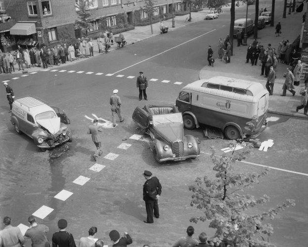 Schwarz-weiß-Szene eines Autounfalls an einer Straßenkreuzung mit mehreren Fahrzeugen, darunter ein Lieferwagen, und einer Gruppe von Menschen in der Nähe, mit Gebäuden, Bäumen und Schildern im Hintergrund.