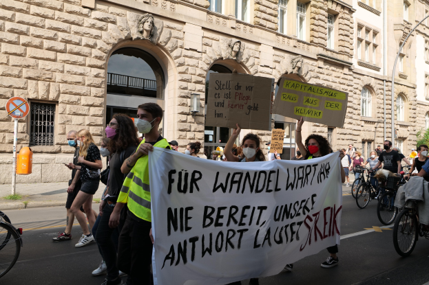 Eine Gruppe von Menschen marschiert bei einer Demonstration durch eine Straße, einige halten Schilder und andere fahren Fahrräder, im Hintergrund ein Gebäude mit Fenstern, Bogengängen, Pfeilern und Skulpturen.