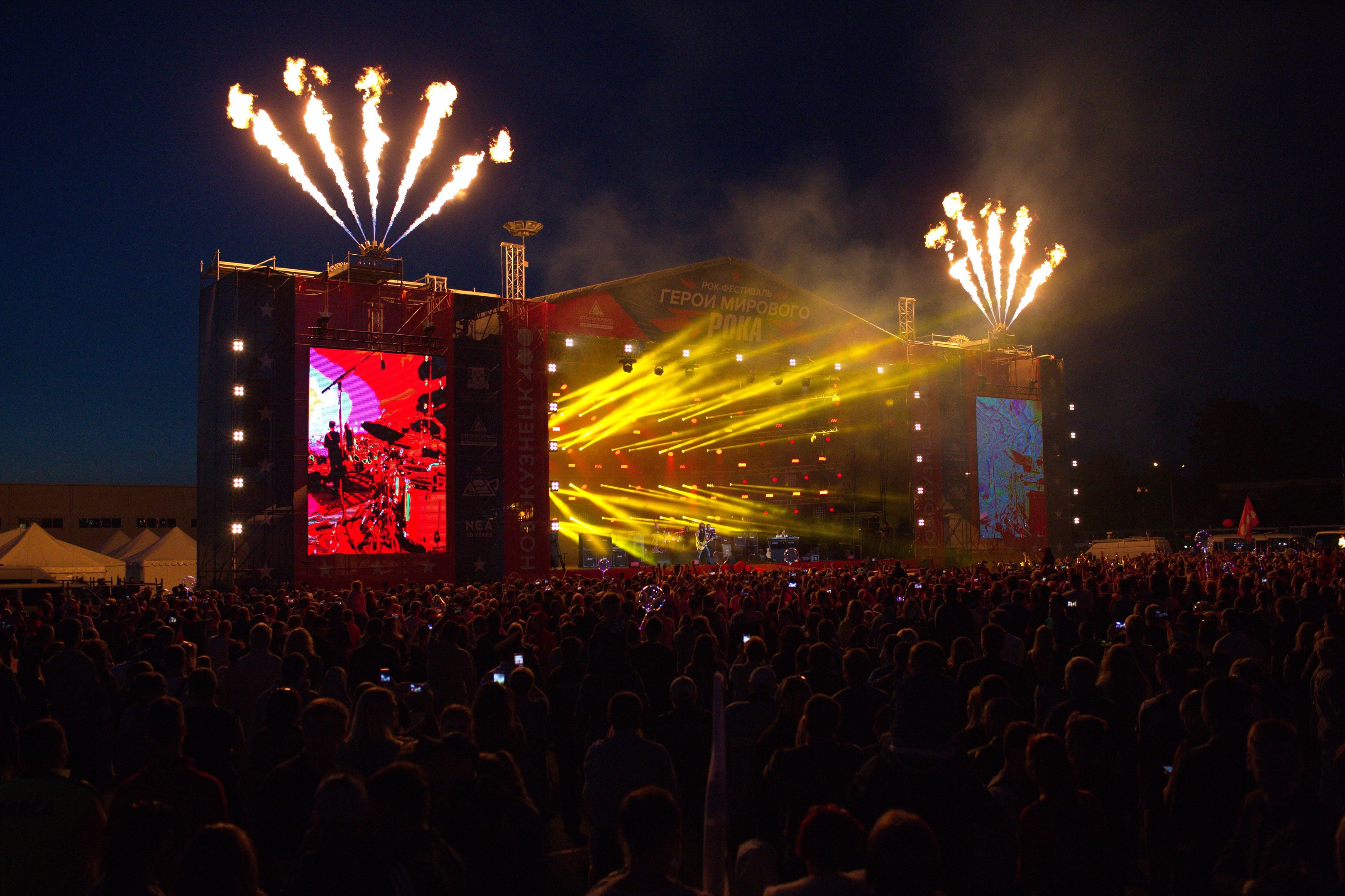 Große Menschenmenge auf einem Musikfestival mit beleuchteter Bühne, auf der Künstler spielen, umgeben von Bannern, Zelten links und Feuerwerk am dunklen Himmel dahinter.