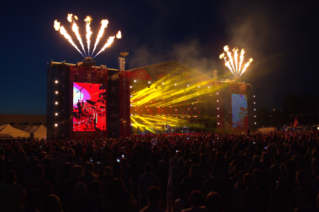 Große Menschenmenge auf einem Musikfestival mit beleuchteter Bühne, auf der Künstler spielen, umgeben von Bannern, Zelten links und Feuerwerk am dunklen Himmel dahinter.