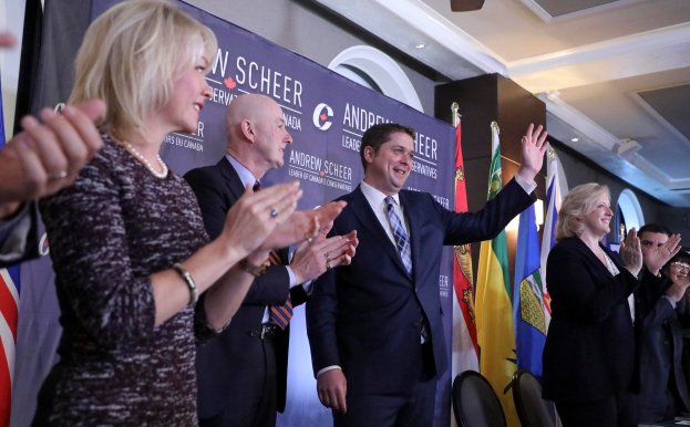 Eine Gruppe von Menschen, die vor Fahnen stehen und jubeln, mit einem Tisch voller Gegenstände rechts daneben, Stühlen, einer Fahne und einer Wand im Hintergrund sowie Lampen und einem Ventilator auf dem Dach