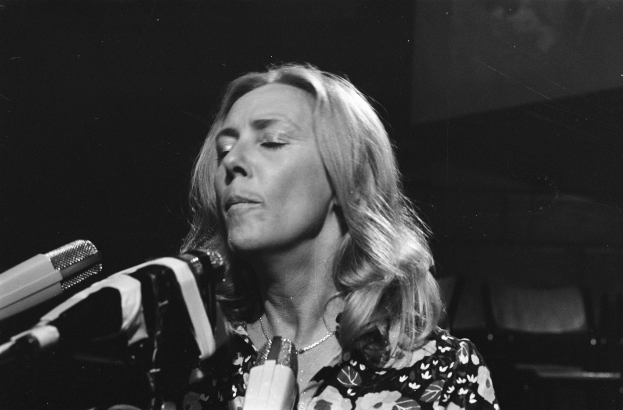 Eine Frau in einem schwarzen und weißen Outfit singt leidenschaftlich in ein Mikrofon, ihre Augen sind geschlossen und ihre Haare sind zu einem klassischen Bob frisiert.