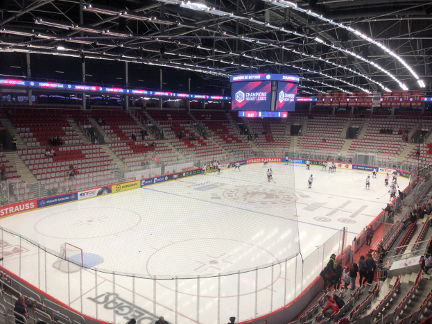 Ein Eishockeyspiel in einer großen Arena zwischen den St. Louis Blues und den Detroit Red Wings, mit Zuschauern auf den Sitzen und Stehplätzen in der Nähe des Spielfelds, umgeben von einem Zaun, einem Bildschirm mit Teamvergleich, Metallrahmen, Deckenleuchten und Dachstruktur.