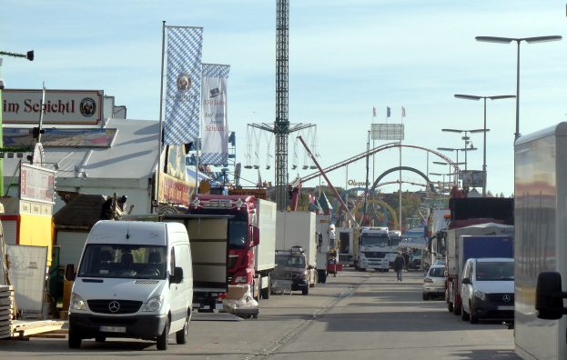 Volles Straßenbild mit Lastwagen, Autos, Fußgängern, Laternen, Bannern, einem Turm, einer Attraktion, Bäumen und einem bewölkten Himmel.