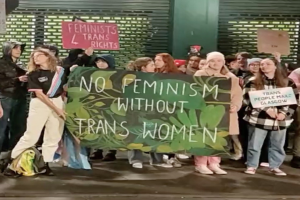 Gruppe von Menschen mit einem Banner "Kein Feminismus ohne Trans-Frauen" und Schildern in einem öffentlichen Raum mit einer Wand und einem Rollladen im Hintergrund.
