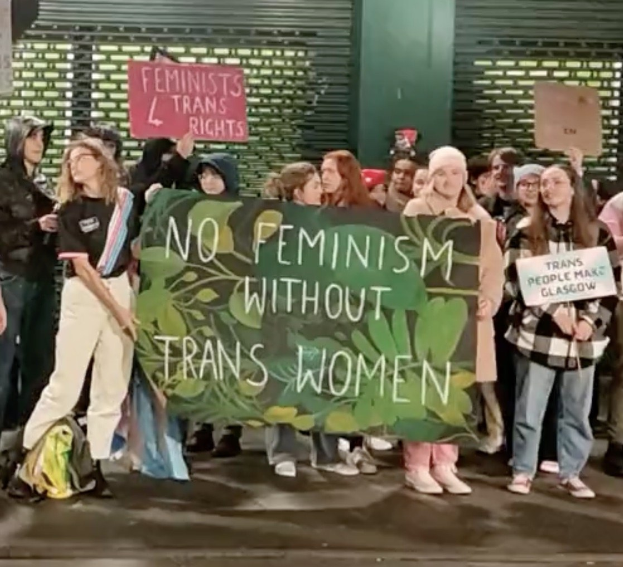 Gruppe von Menschen mit einem Banner "Kein Feminismus ohne Trans-Frauen" und Schildern in einem öffentlichen Raum mit einer Wand und einem Rollladen im Hintergrund.