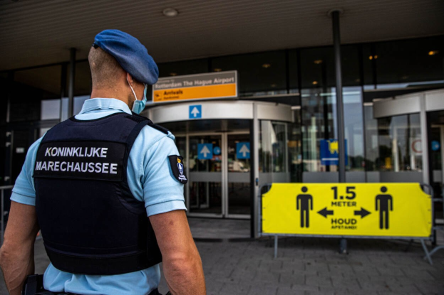 Ein Polizist in einer blauen Uniform und einer Mütze steht vor einem Glasgebäude, trägt eine Maske und hat gelbe und andere Schilder mit Text im Hintergrund.