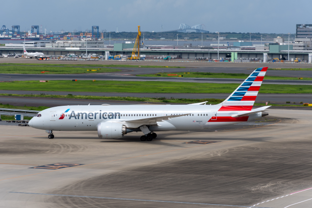 Ein American-Airlines-Flugzeug auf der Rollbahn eines Flughafens, umgeben von Gras, Fahrzeugen, Gebäuden, Masten, Kränen und anderen Gegenständen, mit Bäumen und einem klaren blauen Himmel im Hintergrund.