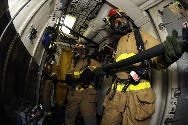Feuerwehrleute in Helmen und Handschuhen mit einer Schlauchleitung in einem Flugzeug mit Ausrüstung und sichtbarem Text im Hintergrund.