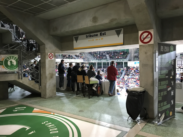 Eine Gruppe von Menschen steht vor einer sitzenden Menge in einem Stadion, mit Schildern, die "Boston Celtics vs New York Jets" lesen, einem Müllcontainer, einer Tafel mit Text, einer Treppe mit Geländern und einem Dach mit Deckenbeleuchtung.