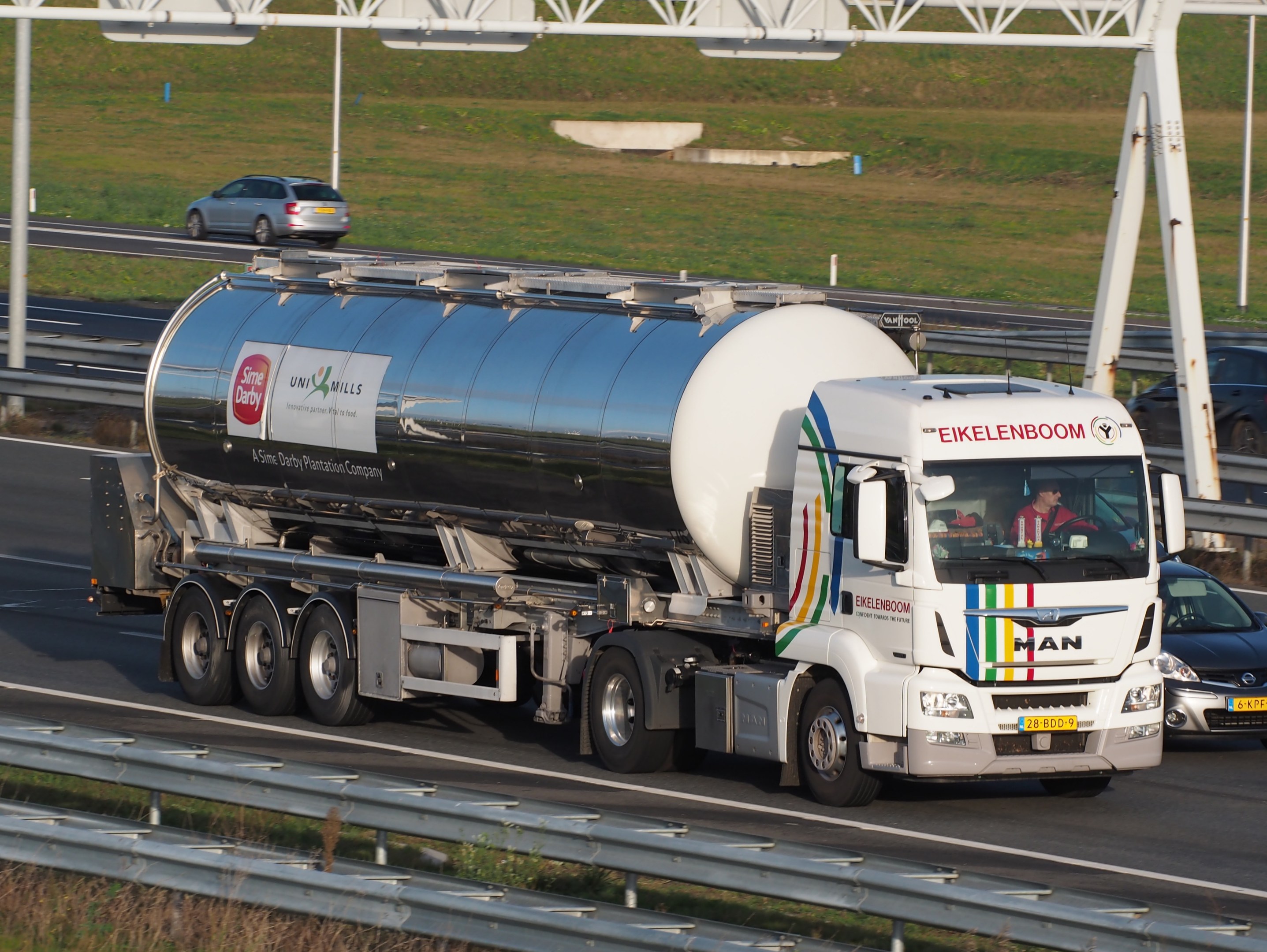 Großer Tanklastzug fährt auf einer Autobahn neben einem Auto, mit sichtbaren Insassen im Truck und grasigem Umfeld einschließlich Stangen und anderen Gegenständen im Hintergrund.