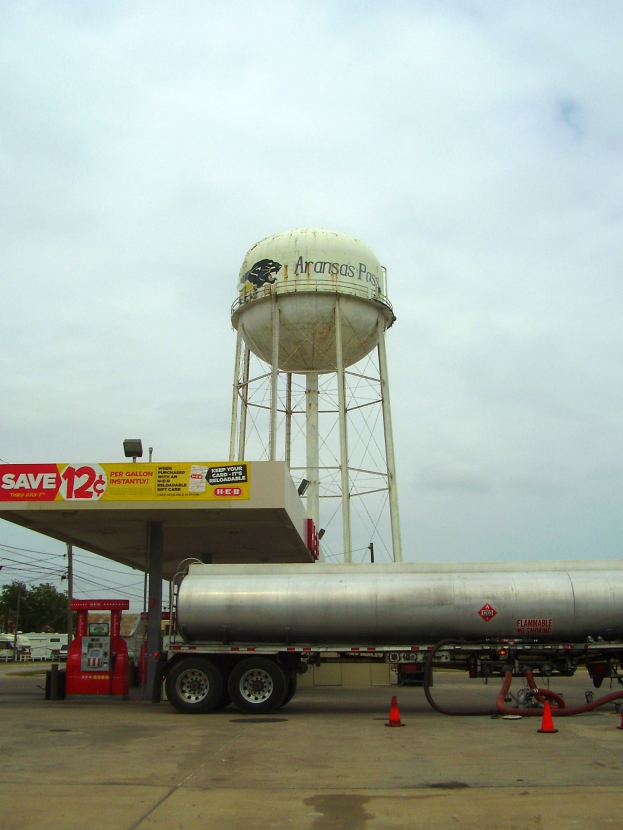 Großer Tanklastzug vor einer Tankstelle mit Zapfsäule, Absperrgittern und anderen Fahrzeugen auf der Straße, mit einem Wasserturm, Bäumen, Masten, Drähten und einem klaren blauen Himmel im Hintergrund.