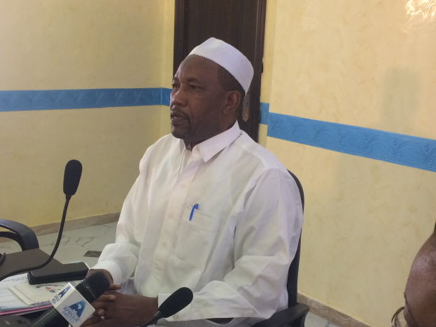 Ein Mann in einem weißen Kleid und Hut sitzt an einem Tisch mit Mikrofonen, spricht, während Papiere und Gegenstände auf dem Tisch zu sehen sind, mit einem anderen Menschen mit Brille rechts von ihm, in einem Raum mit einer Wand und einer Tür dahinter.