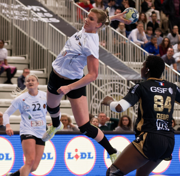 Eine Gruppe von Frauen, die auf einem Platz Handball spielen, mit einem Ball in der Luft, Zuschauern auf Treppen und einem "Handball-Weltmeisterschaft 2019 - Tag 2"-Schild im Hintergrund.