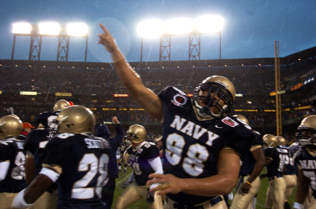 Navy-Football-Spieler in Helmen und Uniformen feiern ein Touchdown auf einem regennassen Rasenfeld, mit Zuschauern und Stadionbeleuchtung im Hintergrund.