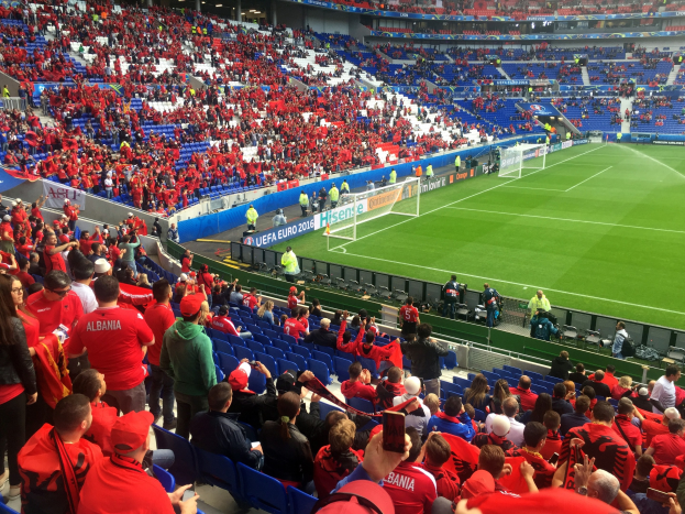 Eine große Menge Menschen in einem Stadion schaut ein Fußballspiel, mit Menschen auf Stühlen und Stehplätzen, Fahnen, einem Torpfosten und Deckenleuchten.