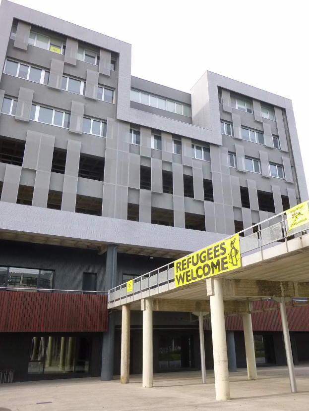 Große Gebäude mit einem "Flütchtlinge Willkommen"-Schild, umgeben von Säulen und einem Geländer, unter einem Himmel-Hintergrund.