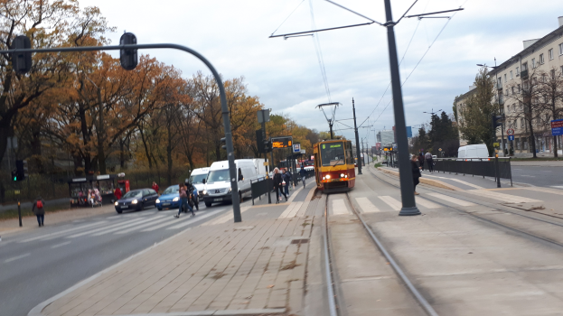 Eine Stadtstraße mit einer gelben Straßenbahn auf den Schienen, Fahrzeugen, Fußgängern, Strommasten mit Leitungen, Verkehrsampeln, Hinweisschildern, Bäumen, Gebäuden mit Fenstern und einem bewölkten Himmel.