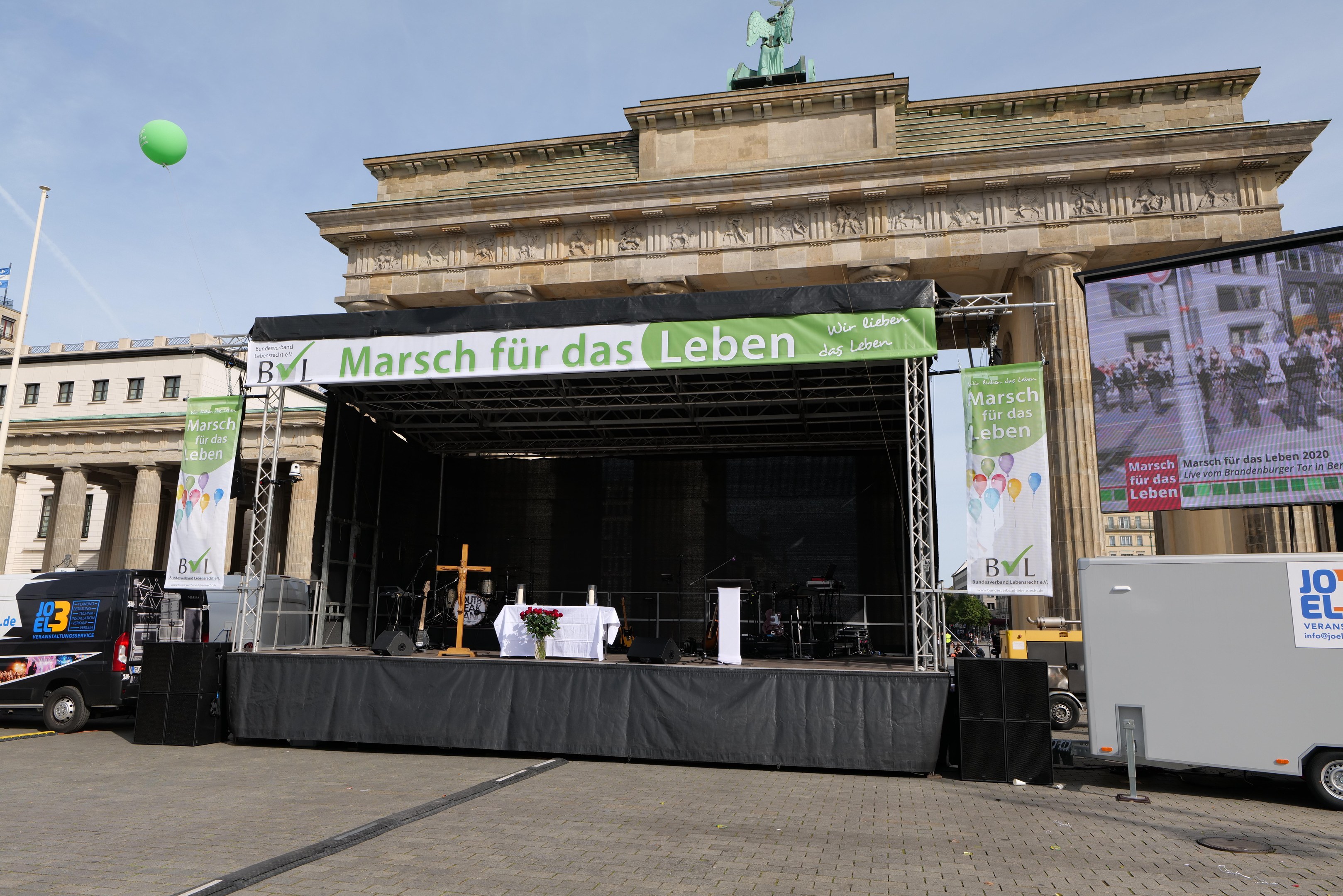 Bühne vor dem Brandenburger Tor mit einem Tisch, Bannern, Rednern, Fahrzeugen, Gebäuden, einer Statue, einer Fahne und Wolken im Hintergrund.