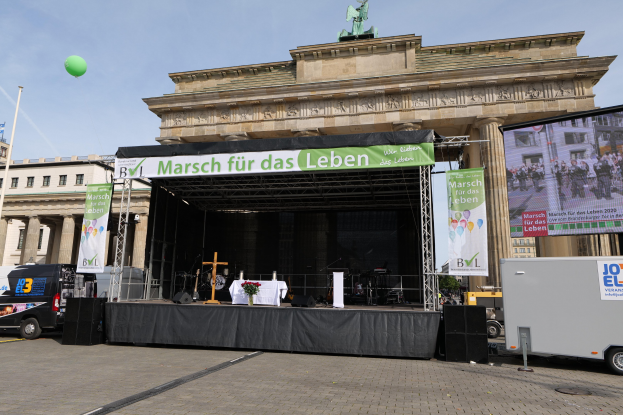 Bühne vor dem Brandenburger Tor mit einem Tisch, Bannern, Rednern, Fahrzeugen, Gebäuden, einer Statue, einer Fahne und Wolken im Hintergrund.