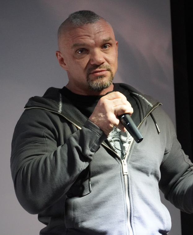 Ein Mann in einer schwarzen Kapuzenjacke und einem schwarzen Anzug mit einem wei├čen Hemd, der ein Mikrofon halt, steht selbstbewusst vor einer Wand und scheint ein Komiker mit zurückgek├Ąmmten Haaren zu sein.