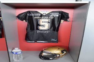 Ein Fußballtrikot und Helm hängen an einer Spindtür, mit einer Wasserflasche auf dem Regal darunter.