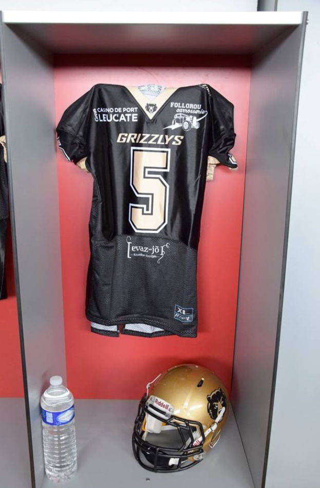 Ein Fußballtrikot und Helm hängen an einer Spindtür, mit einer Wasserflasche auf dem Regal darunter.