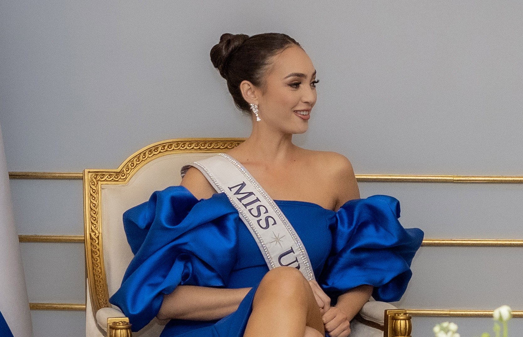 Eine Frau in einem blauen Kleid trägt eine 'Miss Universe 2019'-Schärpe und sitzt lächelnd auf einem Stuhl, umgeben von Blumen und einer Wand im Hintergrund.