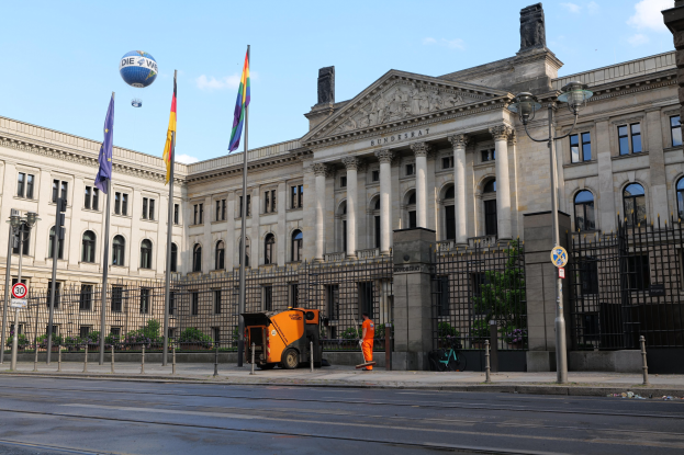 Großes modernes Gebäude mit vielen Fenstern, als Bundestag in Berlin identifiziert, umgeben von Straßeninfrastruktur, Fahrzeugen, Fußgängern und Bäumen unter einem bewölkten Himmel.