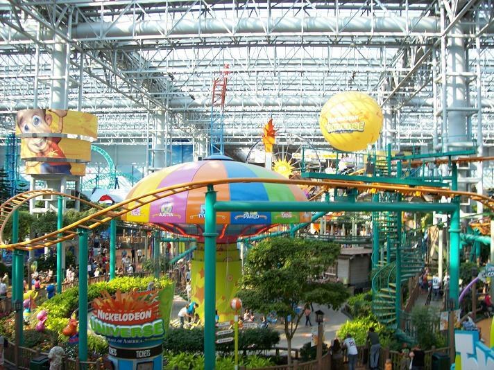 Ein großer Indoor-Wasserpark mit einer Achterbahn, umgeben von Menschen, Bäumen, Pflanzen und Deckenbeleuchtung, die eine lebendige Atmosphäre schaffen.