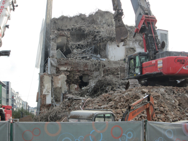Ein Kran im Vordergrund zerstört ein Gebäude, umgeben von anderen Kränen, Fahrzeugen, Felsen und einer Fahne mit Text, mit Gebäuden, Bäumen und Himmel im Hintergrund.