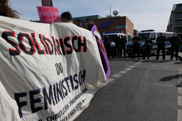 Gruppe von Menschen, die eine Straße entlanggehen und eine 'Solidarität und Feminismus'-Fahne tragen, mit parkenden Fahrzeugen, Gebäuden, einer Schüsselantenne und einem klaren blauen Himmel im Hintergrund.