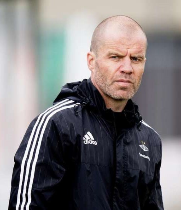 Kahlköpfiger Trainer in marineblauer Jacke mit weißen Streifen und weißem Kragen steht auf einem Fussballfeld mit verschränkten Händen, ernsthafter Gesichtsausdruck, unscharfer Hintergrund.