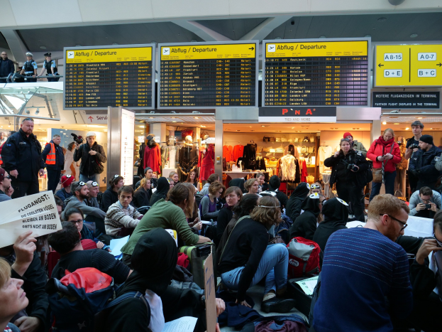 Eine große Gruppe von Menschen an einem Flughafen, einige sitzen mit Taschen und Papieren, andere stehen, mit Schildern, Schaufensterpuppen in Kleidern und Deckenlampen im Hintergrund, was auf eine Protestveranstaltung hindeutet.
