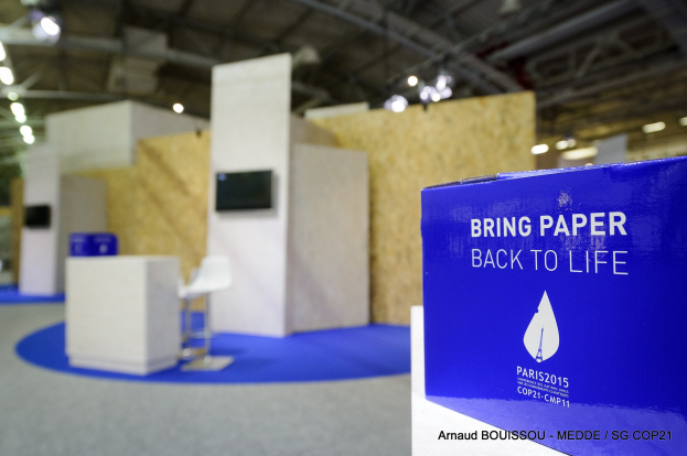 Messestand mit einem blauen und weißen "Bring Paper Back to Life"-Schild auf der rechten Seite, das vor einem Hintergrund aus leicht unscharfen Stühlen, Tischen, einer Wandfernseher und Deckenleuchten zu sehen ist.