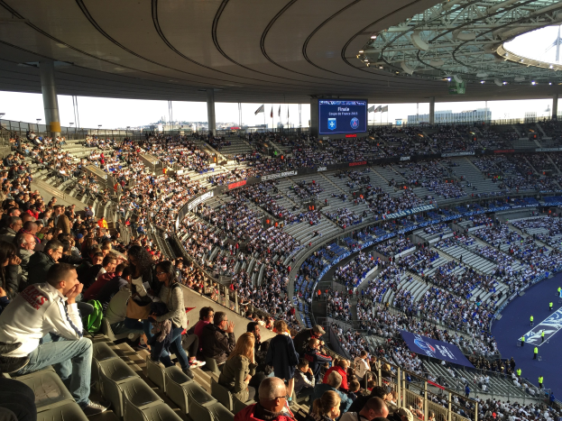 Große Menschenmenge in einem Stadion bei einem Fussballspiel, mit einer Bühne rechts, Fahnen, Masten, einem Bildschirm und der Allianz Arena in München, Deutschland im Hintergrund.