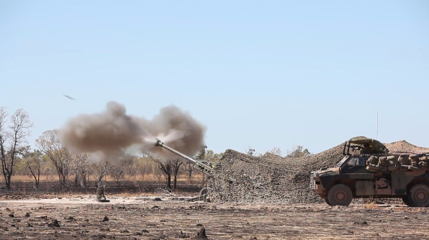 Militärfahrzeug mit Rauchentwicklung auf einer Schotterstraße unterwegs, mit einem Soldaten an Bord und Bäumen im Hintergrund bei klarem blauem Himmel.