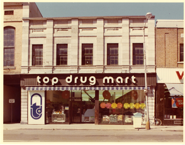Ein Drogenladen an einer Straßenecke mit einem Gebäude, einem Schild, einem Straßenpfosten, einem geparkten Fahrrad, einem Müllcontainer und einem bewölkten Himmel.