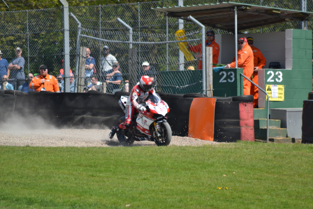 Eine Person mit Helm und Handschuhen fährt ein Motorrad auf einem grasbewachsenen Feld, mit Zuschauern, Absperrungen, Treppen und einem Schuppen im Hintergrund.