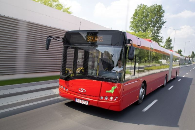 Ein roter Bus fährt auf einer Straße neben einem Gebäude, mit einer Person im Inneren, neben einer Wand, Bäumen, Laternen und einem bewölkten Himmel.