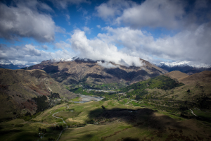 Eine Panoramansicht von einem Berggipfel in Queenstown, Neuseeland, zeigt eine gewundene Straße, grünes Gras, Bäume und einen Himmel voller weißer, flauschiger Wolken.