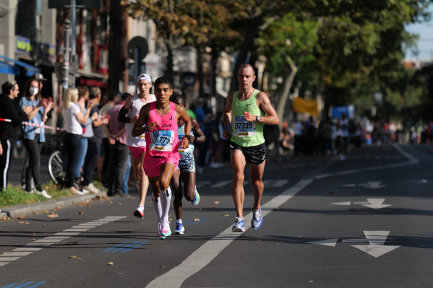 Gruppe von Menschen beim Stadtmarathon, mit Zuschauern auf der linken Seite, unscharfen städtischen Hintergrund mit Bäumen, Gebäuden und einem Fahrrad.