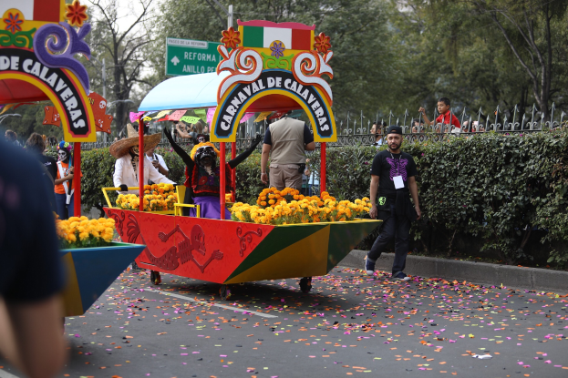 Eine Gruppe von Menschen auf einem blumengeschmückten Festwagen bei einem Umzug, die verschiedene bunte Kleider und Hüte tragen, mit Schildern, einem Metallzaun, Bäumen und einem bewölkten Himmel im Hintergrund.