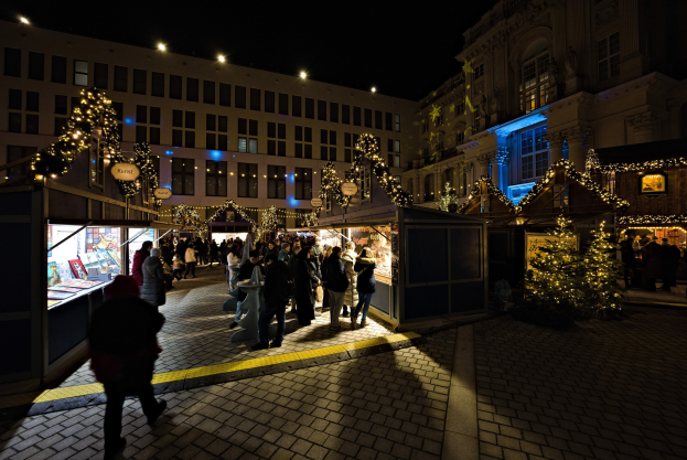 Ein belebter Weihnachtsmarkt in der Nacht in einer Stadt, mit Menschen um geschmückte Stände und beleuchtete Gebäude im Hintergrund.